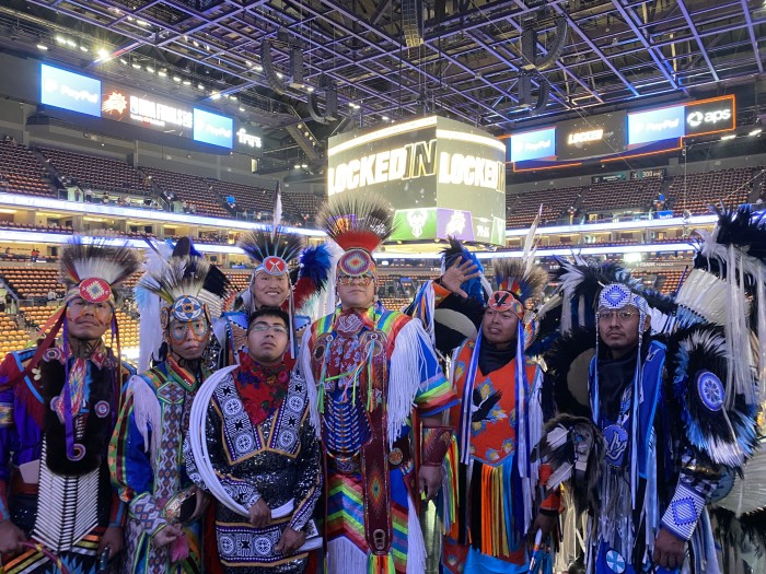 Native men in colorful feather headresses and beaded outfits pose inside a big event hall.